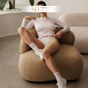 A slim young woman is sitting casually on a sofa with one leg bent. She is wearing a sporty outfit consisting of a slightly transparent long-sleeve top, white bottoms, and white sneakers.