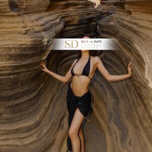 Eine zierliche brünette Frau steht zwischen zwei schmalen Felsklippen. Sie trägt einen knappen schwarzen Bikini sowie einen schwarzen Wickelrock und blickt in die Kamera.