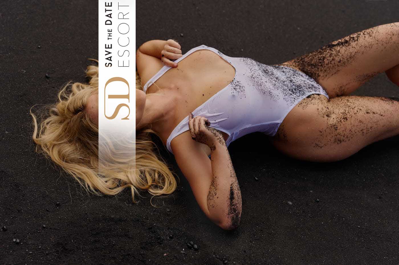 A sporty blonde woman is lying on her back on black sand. She is wearing a white swimsuit with a deep neckline and smiling sweetly into the camera.