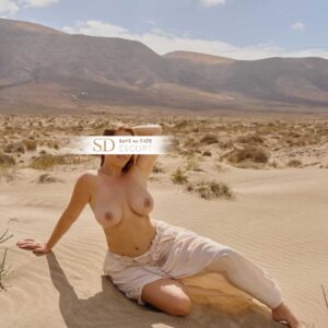 Eine vollbusige, kurvige junge Frau sitzt posierend im Sand, streicht sich mit der Hand durch die Haare und trägt dabei nur eine Hose.